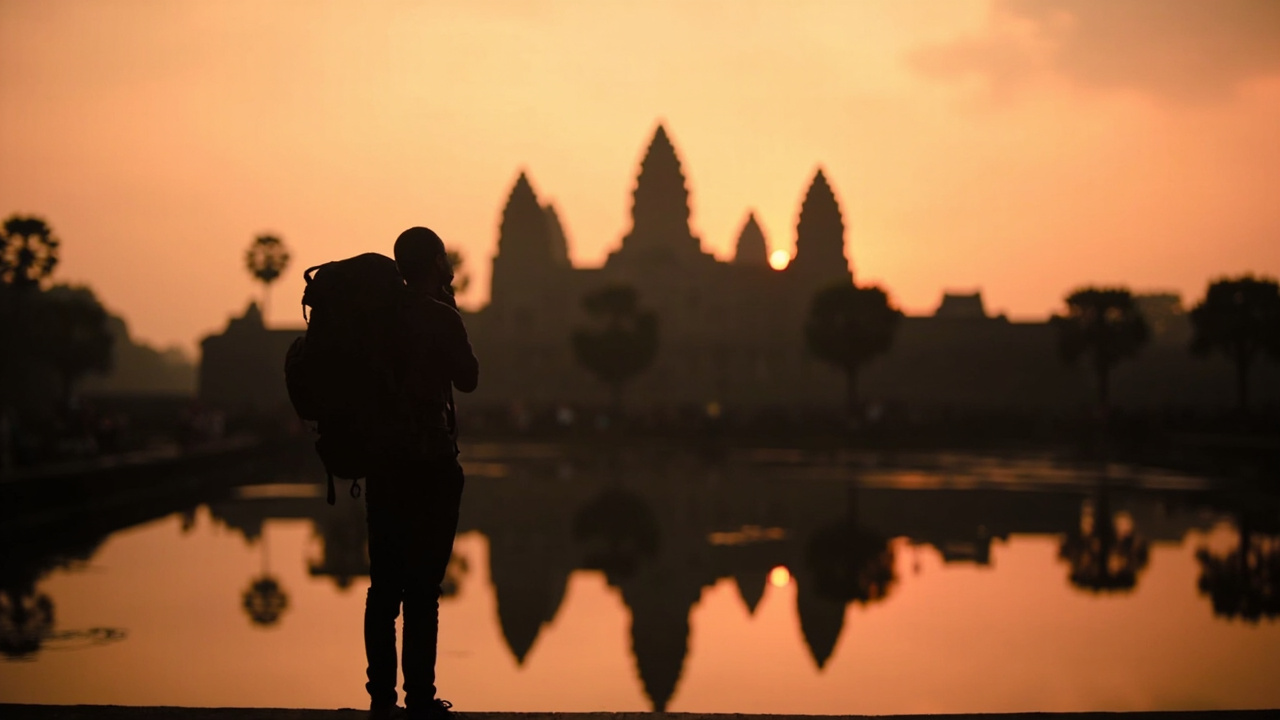 Silhouette of the central towers of Angkor Wat at sunrise, reflecting pond in the foreground, an Australian traveller in soft silhouette to one side watching the sky turn warm gold and pink